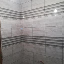 PWO - Ściana łazienki wyłożona szarymi płytkami imitującymi drewno z poziomymi pasami dekoracyjnymi, widoczny fragment instalacji wodnej.