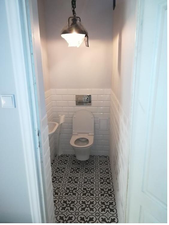 Mała łazienka po remoncie z toaletą, umywalką, białymi płytkami na ścianach i wzorzystą czarno-białą podłogą, oświetlona lampą wiszącą w stylu industrialnym.
