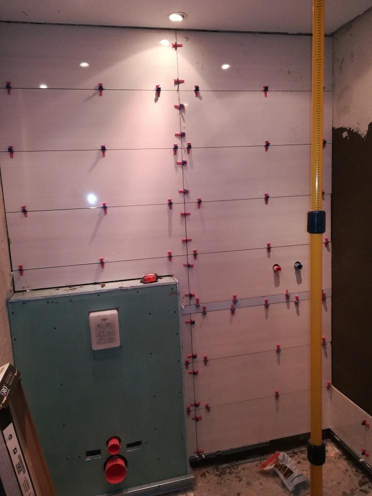 Ściana łazienki w trakcie układania jasnych, prostokątnych płytek, z użyciem czerwonych krzyżyków dystansowych i poziomicy; widoczne elementy instalacji wodnej i obudowa z płyty gipsowej.