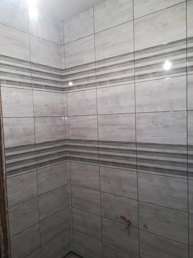 Ściana łazienki wyłożona szarymi płytkami imitującymi drewno z poziomymi pasami dekoracyjnymi, widoczny fragment instalacji wodnej.