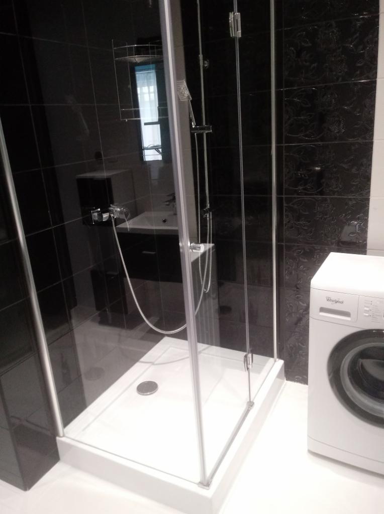 Nowoczesna kabina prysznicowa z brodzikiem, czarne płytki z fakturą, umywalka z szafką i pralka w tle, widok z boku.