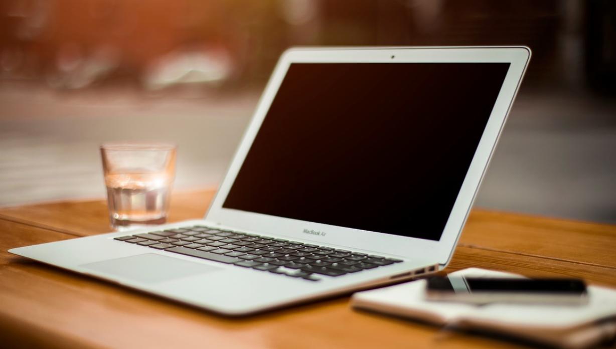 Laptop MacBook Air z pustym ekranem, szklanka wody i smartfon na notatniku na drewnianym stole w kawiarni.