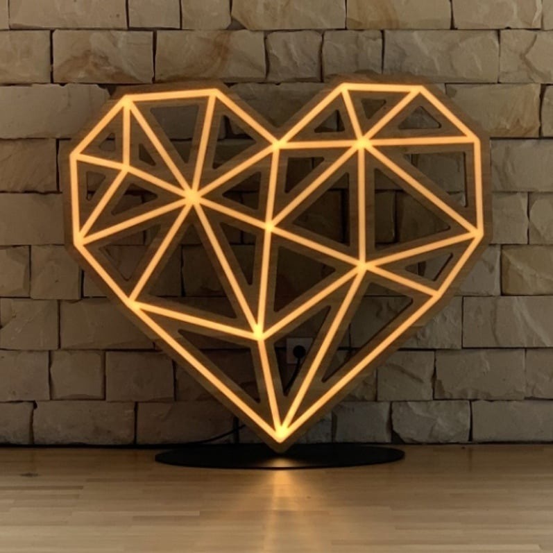 Dekoracyjne serce z drewnianą ramą i wewnętrznym oświetleniem LED w geometrycznym wzorze trójkątów, ustawione na podłodze z jasnego drewna na tle ściany z kamienia dekoracyjnego.