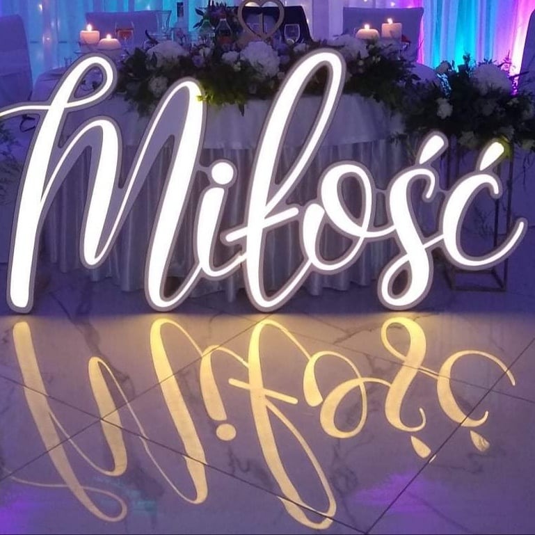 Podświetlany napis 'Miłość' w ozdobnej czcionce, ustawiony przed stołem weselnym z kwiatami i świecami, widoczne odbicie napisu na błyszczącej podłodze.