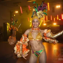 Uśmiechnięta tancerka w bogatym, karnawałowym stroju z piórami i cekinami, występuje na scenie podczas imprezy, w tle widoczne oświetlenie i publiczność.