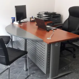 Biuro z biurkiem o blacie z drewna i szarej płyty, czarnymi krzesłami, komputerem, laptopem i drukarką, na podłodze ciemne płytki imitujące marmur.