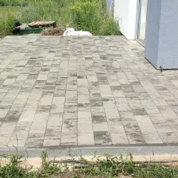 Ułożona kostka brukowa w odcieniach szarości z widoczną fakturą, przylegająca do białego budynku, z paletą i workiem w tle, w otoczeniu trawy.