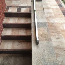 Kamienne schody zewnętrzne z brązowymi stopniami i poziomnicą, przylegające do ceglanej ściany i wyłożonego kostką brukową chodnika.