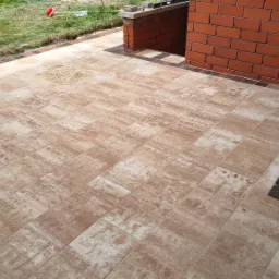 Nowo ułożony taras z beżowo-brązowej kostki brukowej, wykończony ciemniejszą obwódką, przylegający do ceglanego murku i ściany budynku z zewnętrznym kranem.