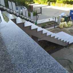 Brukki - Zewnętrzne schody z szarego kamienia z metalową barierką, widok z góry, teren wokół w trakcie prac wykończeniowych, widoczny wąż ogrodowy i kontener na gruz.