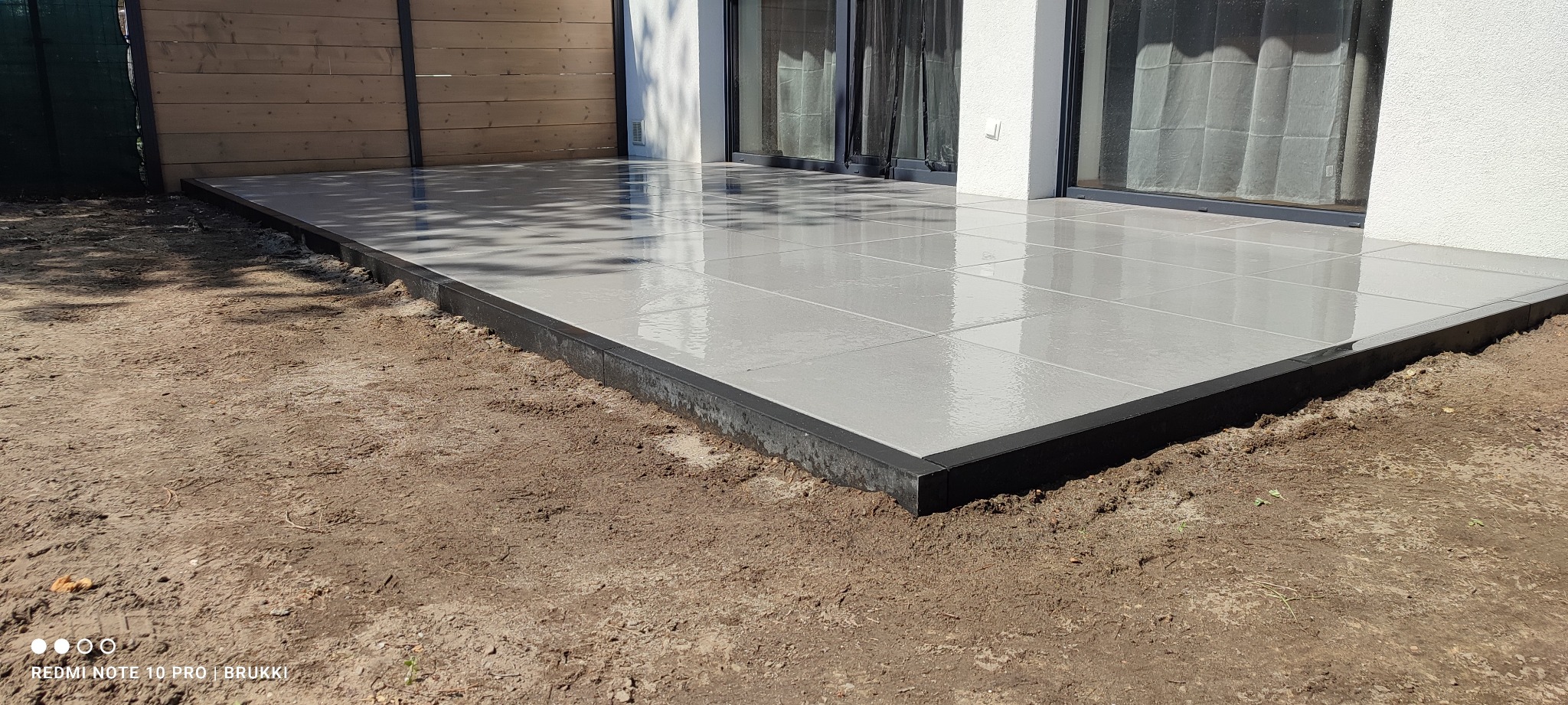 Nowoczesny taras z szarych płyt kamiennych, wykończony czarnym obrzeżem, przylegający do budynku z dużymi oknami tarasowymi, widok na świeżo przygotowany teren.