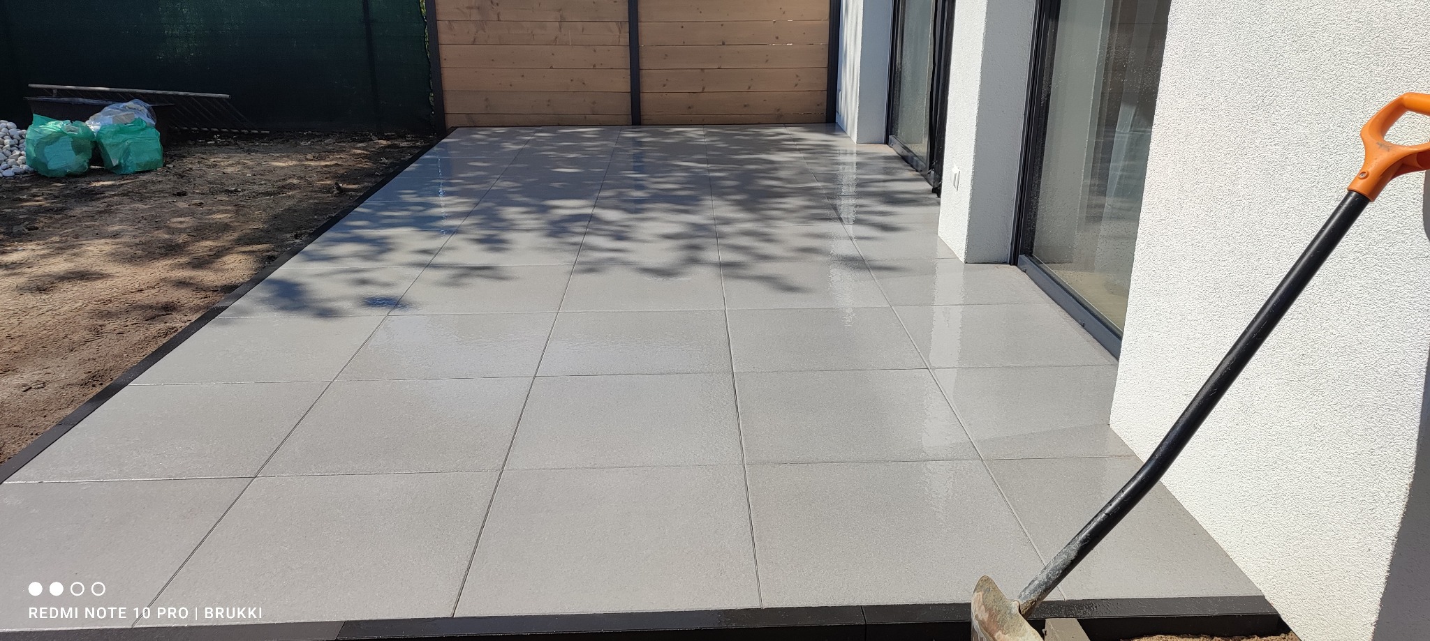 Nowoczesny taras wyłożony szarymi płytami z widoczną łopatą opartą o ścianę budynku, obok przesuwne drzwi tarasowe i drewniane ogrodzenie w tle.