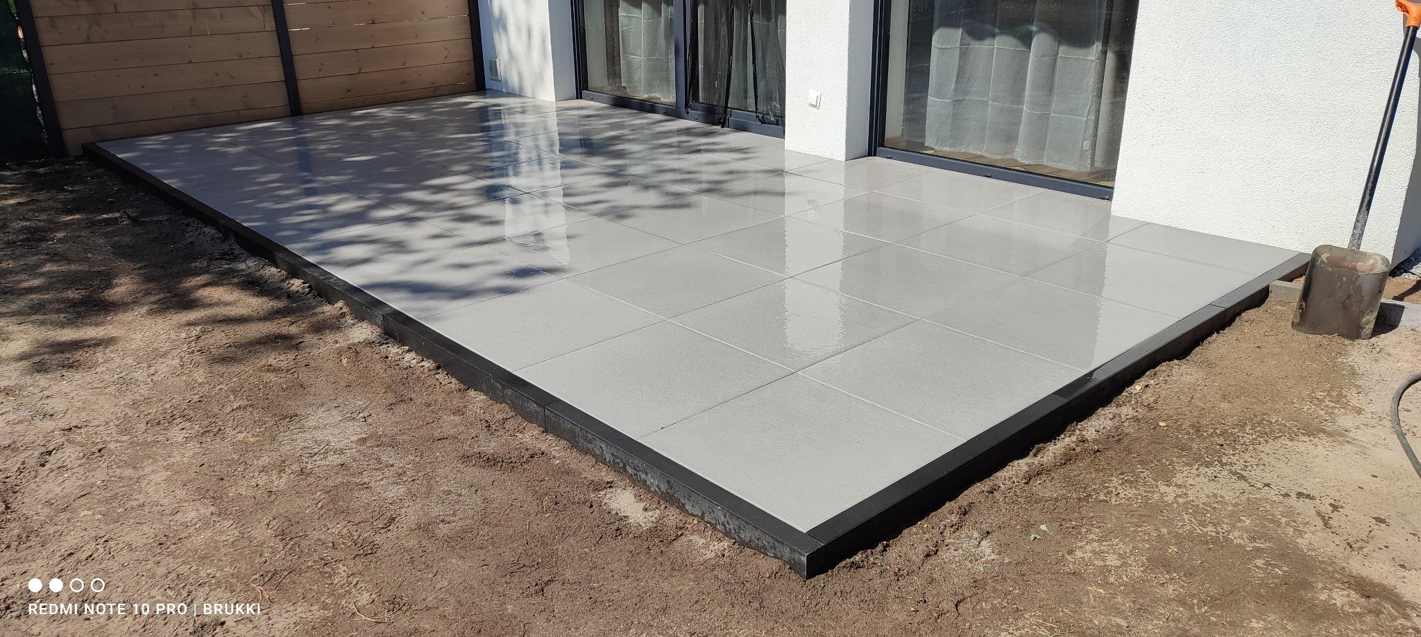 Nowoczesny taras wyłożony jasnoszarą płytką ceramiczną, obrzeża z czarnego kamienia. Na obrzeżach widoczna goła ziemia, w tle budynek z dużymi oknami tarasowymi i drewnianym ogrodzeniem.