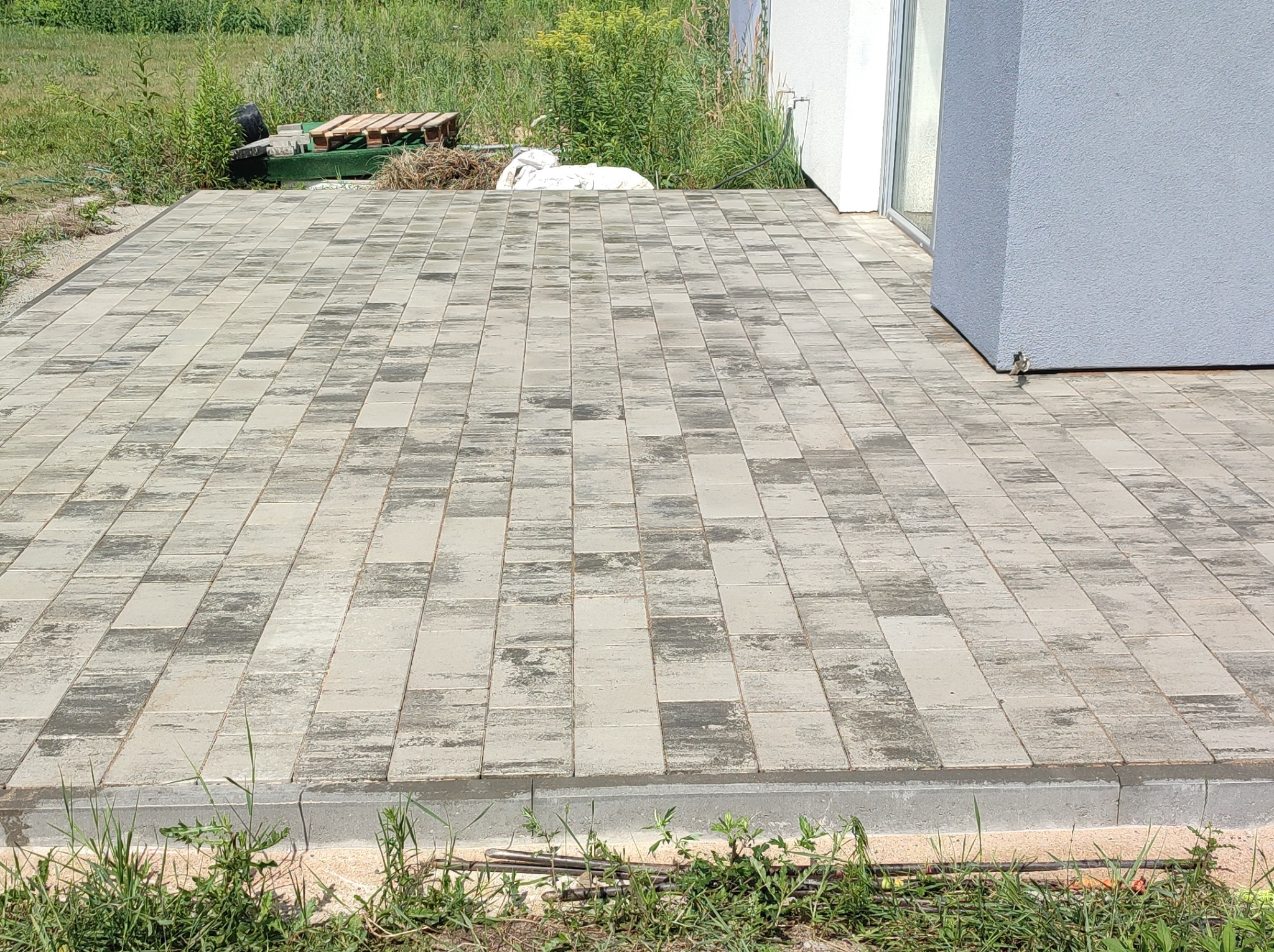 Ułożona kostka brukowa w odcieniach szarości z widoczną fakturą, przylegająca do białego budynku, z paletą i workiem w tle, w otoczeniu trawy.