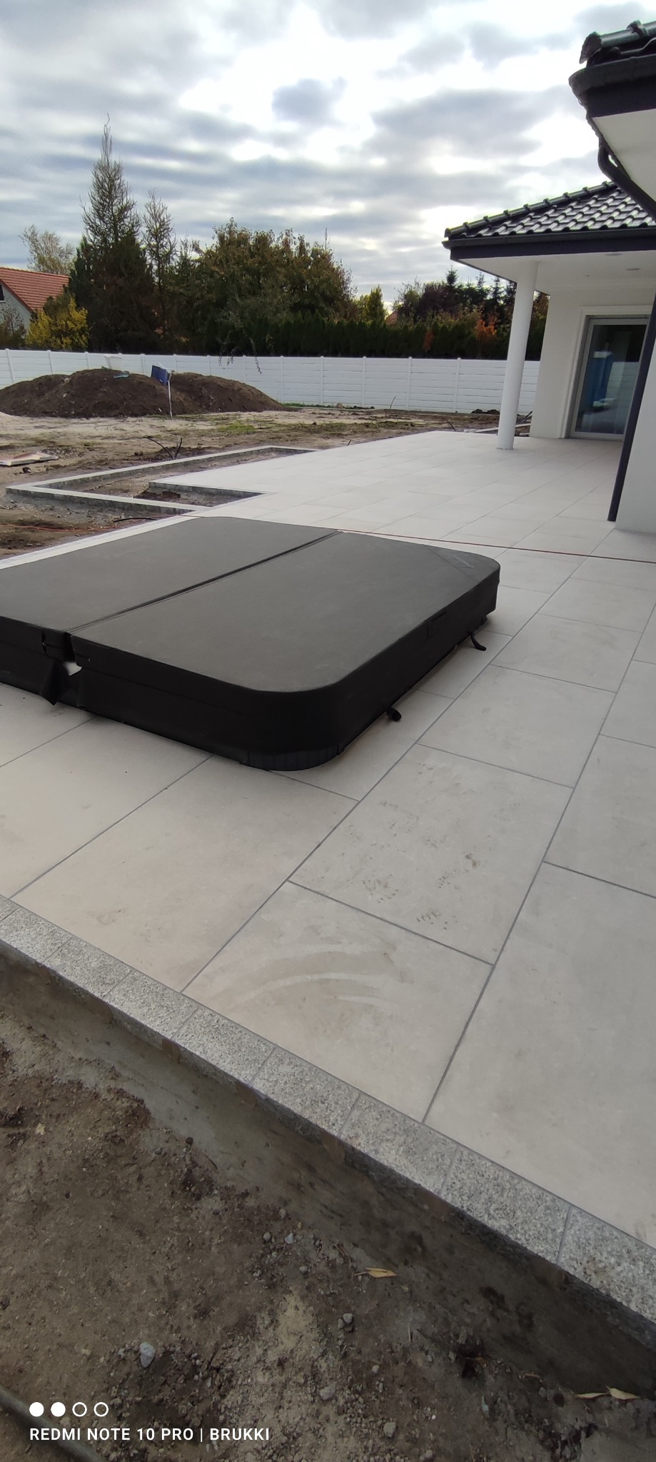 Jasne płytki tarasowe ułożone wokół wanny jacuzzi z czarną pokrywą, widoczne przygotowania do dalszych prac ziemnych w tle.