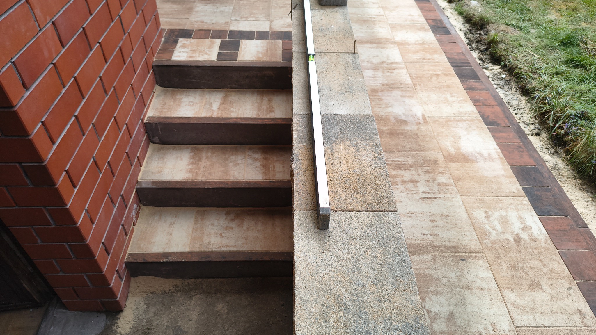 Kamienne schody zewnętrzne z brązowymi stopniami i poziomnicą, przylegające do ceglanej ściany i wyłożonego kostką brukową chodnika.