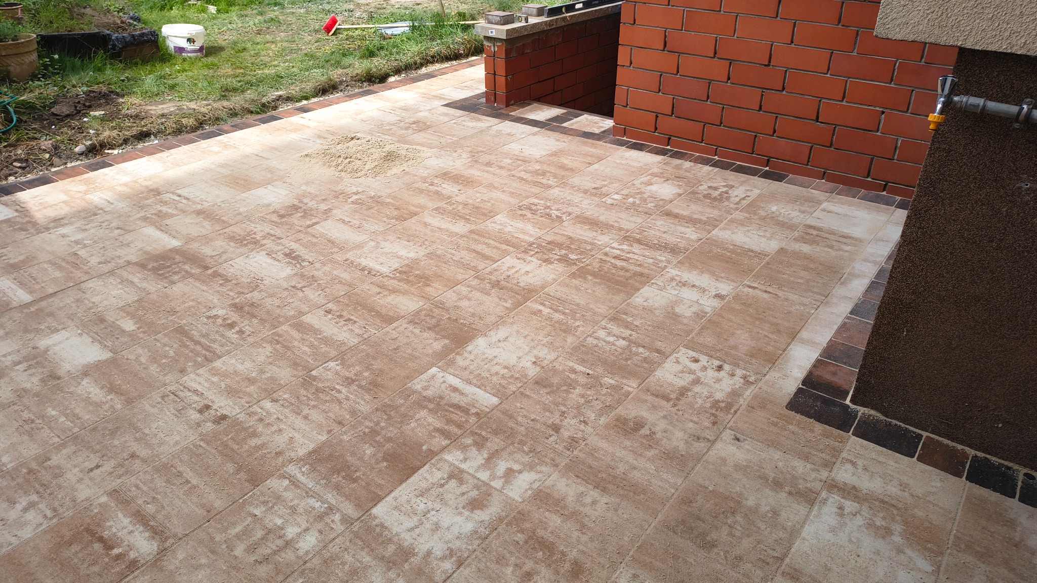 Nowo ułożony taras z beżowo-brązowej kostki brukowej, wykończony ciemniejszą obwódką, przylegający do ceglanego murku i ściany budynku z zewnętrznym kranem.
