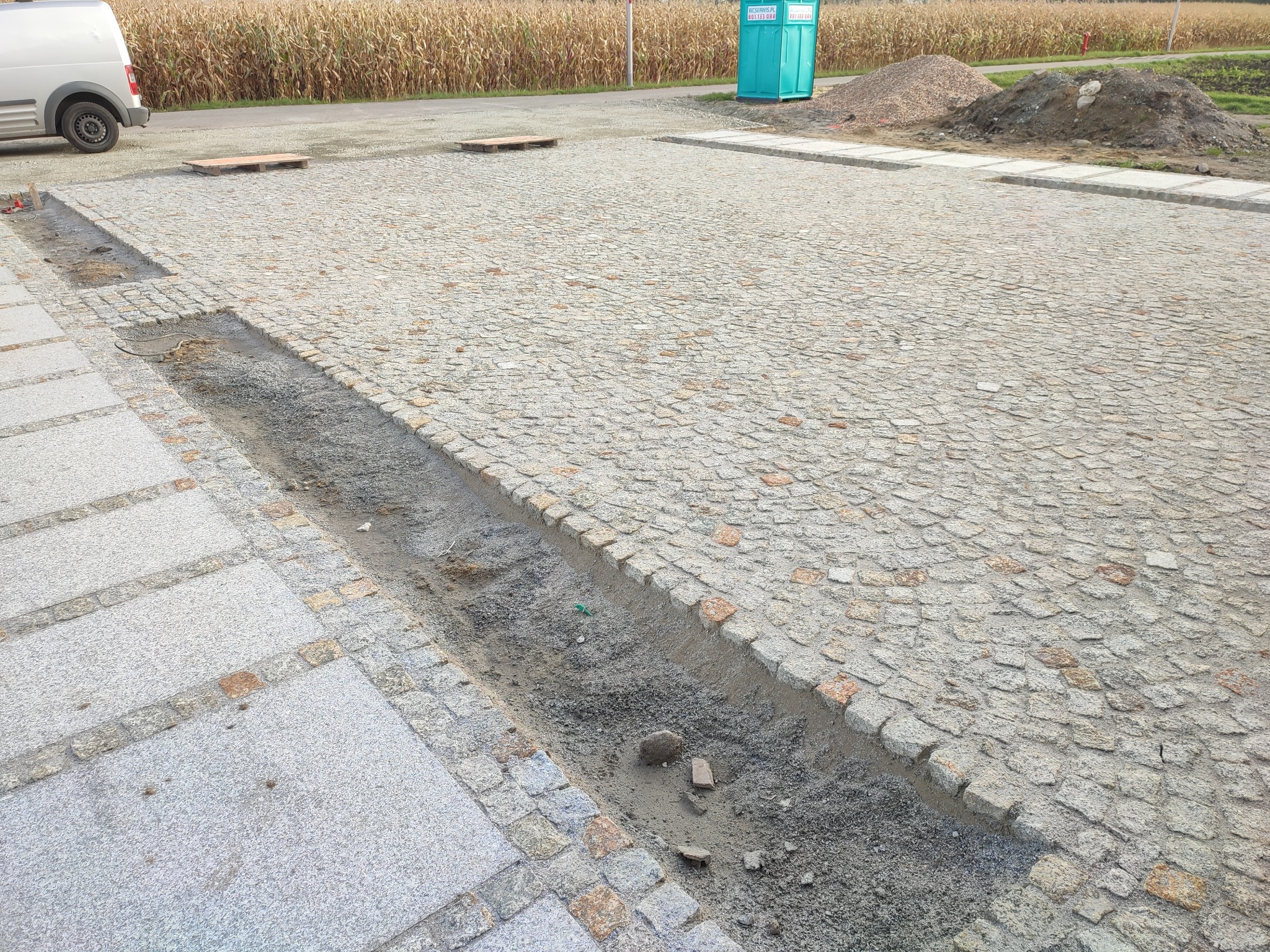 Fragment powierzchni z jasnoszarej kostki granitowej, z widocznym wykopem wzdłuż krawędzi, przygotowanym pod dalsze prace. W tle widoczne pole kukurydzy i przenośna toaleta.