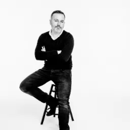 Czarno-białe portretowe ujęcie mężczyzny z brodą, siedzącego na stołku z założonymi rękami na białym tle.