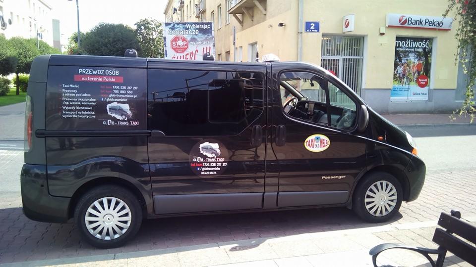 Czarny bus z naklejkami informującymi o świadczeniu usług przewozu osób Taxi Mielec, zaparkowany na chodniku w miejskiej scenerii, widoczny plakat antynarkotykowy i szyld Banku Pekao w tle.