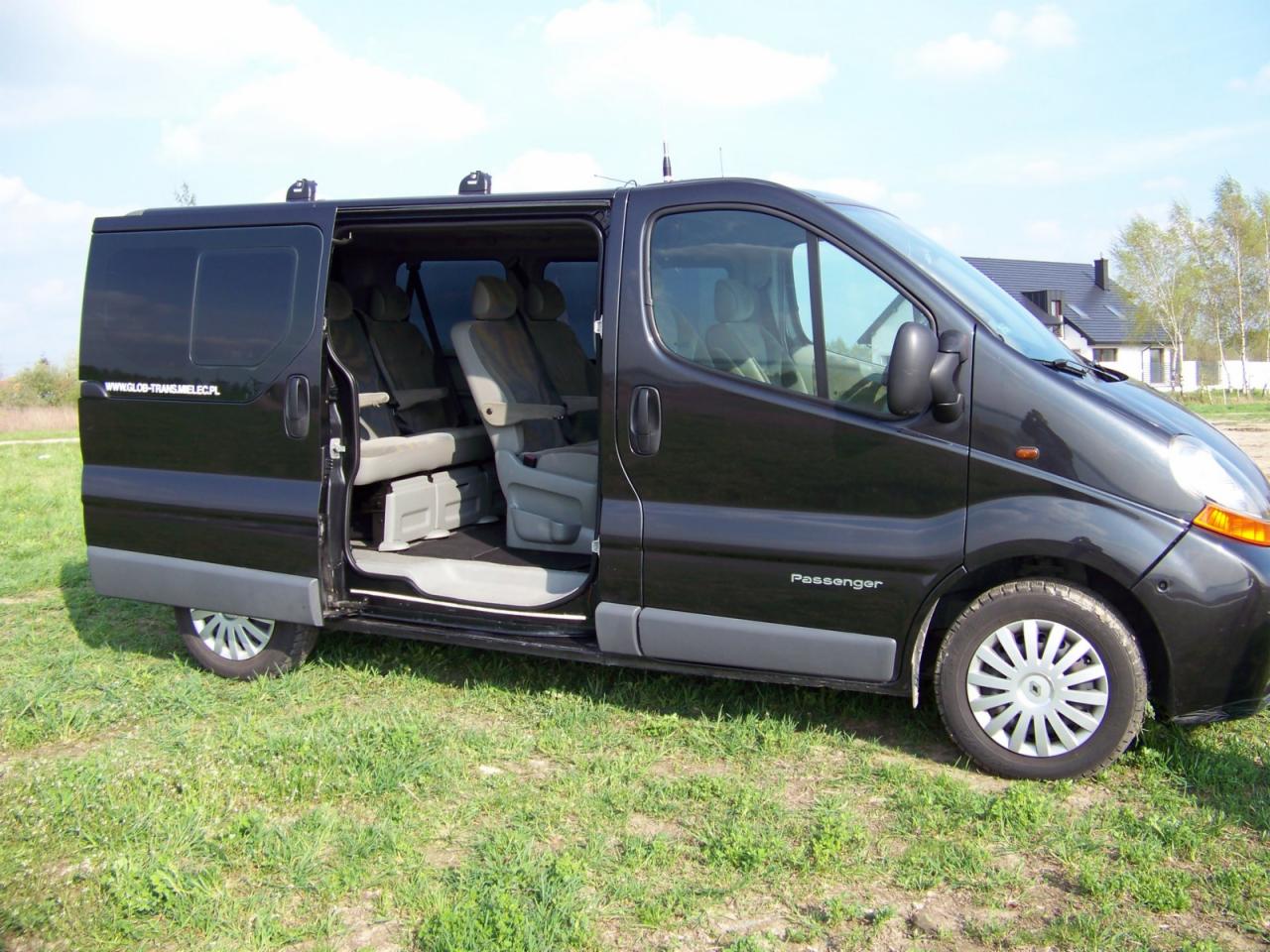 Czarny van pasażerski z otwartymi drzwiami bocznymi, widoczne szare fotele wewnątrz, zaparkowany na trawie przed domem.