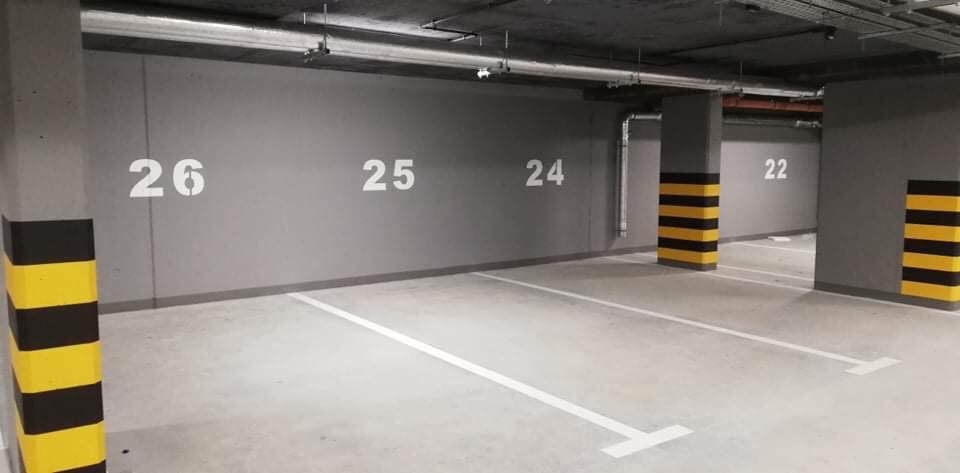 Oznakowane miejsca parkingowe w garażu podziemnym, numery 22, 24, 25 i 26 na szarej ścianie, słupy zabezpieczone pasami żółto-czarnymi.