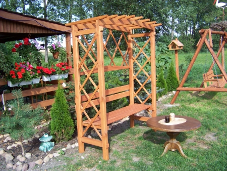 Drewniana pergola z ławką w ogrodzie, obok stolik kawowy i huśtawka, w tle altana z kwiatami.