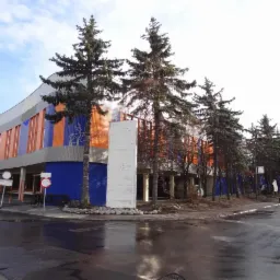 Nowoczesny budynek z pomarańczowo-niebieską elewacją, widziany z rogu ulicy po deszczu, z wysokimi drzewami iglastymi na pierwszym planie.