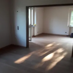 Jasne panele podłogowe w pustym pokoju z białymi ścianami, widoczne przez drewniane futryny drzwi, wpadające promienie słońca