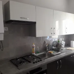 Nowoczesna kuchnia z białymi szafkami, szarym blatem i płytą gazową, widoczny bojler gazowy po lewej stronie, naczynia w suszarce nad zlewem.