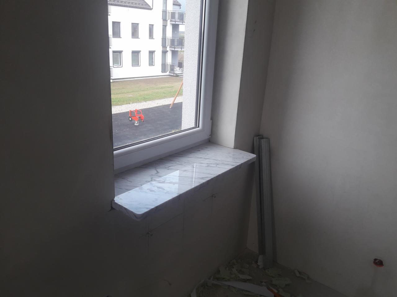 Marmurowy parapet wewnętrzny zamontowany w oknie PCV, widoczne ślady po kleju i ołówku na ścianie, za oknem plac zabaw z czerwonym traktorkiem.