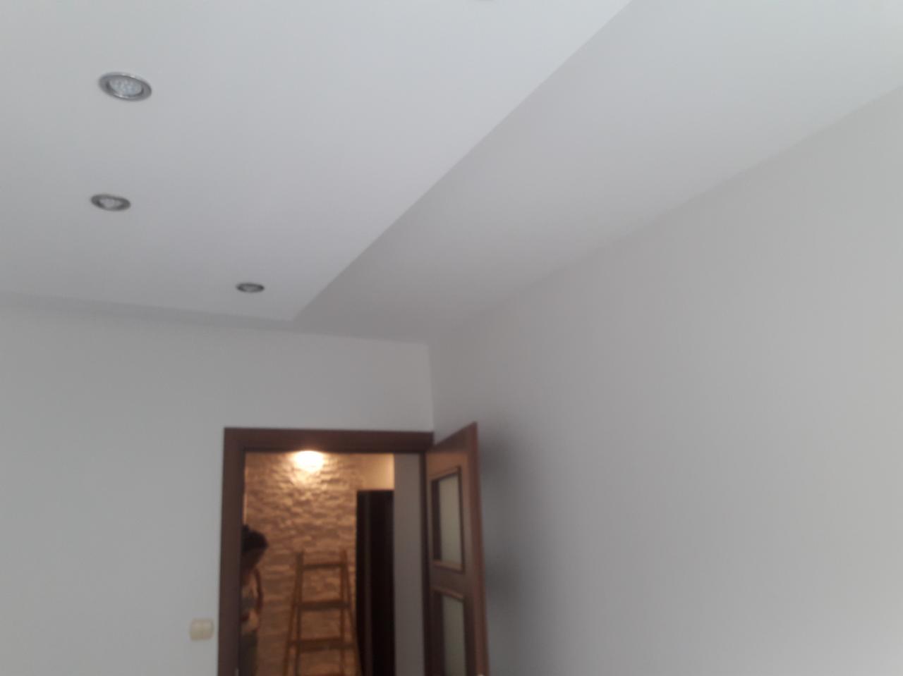 Wnętrze po remoncie: gładkie, białe ściany i sufit z trzema wpuszczonymi punktami świetlnymi; widoczne drzwi z ościeżnicą oraz drabina w tle, sugerujące trwające prace wykończeniowe.
