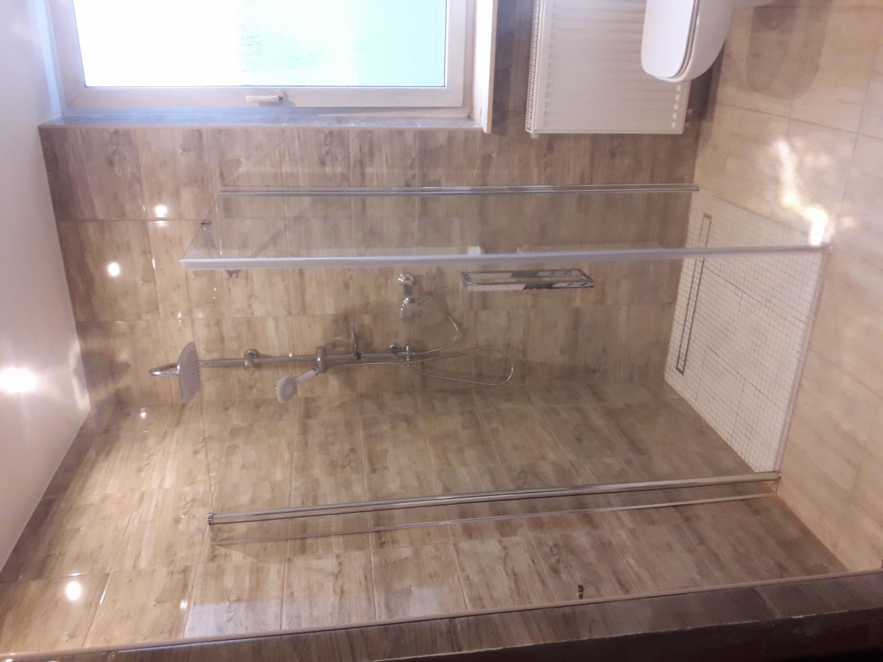 Nowoczesna kabina prysznicowa z deszczownicą i baterią termostatyczną, obłożona płytkami imitującymi drewno, z białymi płytkami na podłodze brodzika i widocznym oknem w górnej części.