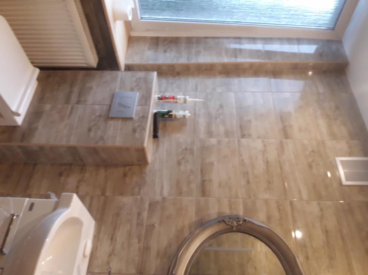 Wykończona łazienka z beżowymi płytkami imitującymi drewno, widoczne: toaleta, umywalka, lustro w ozdobnej ramie, okno, grzejnik, wentylacja, oraz dwie tuby z silikonem i przyłącze hydrauliczne.