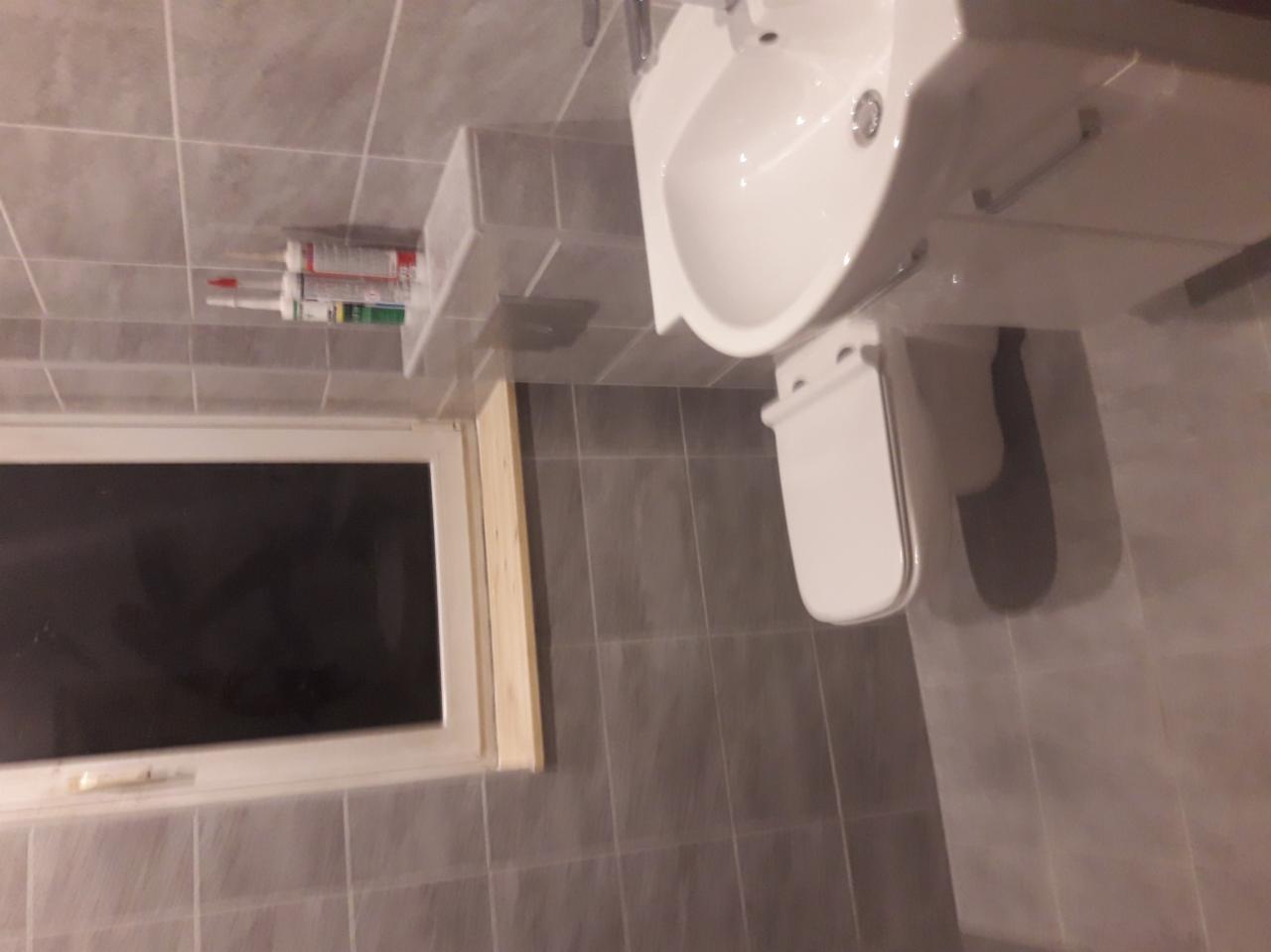 Ukończona łazienka z szarymi płytkami, białą umywalką i toaletą, widoczne tuby z silikonem na półce nad oknem.
