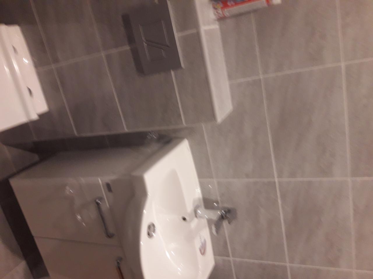 Nowa, biała umywalka z szafką w szarej łazience z płytkami, widoczne elementy instalacji i armatura, ujęcie z góry.