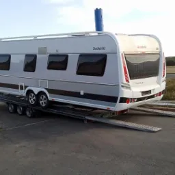 Przyczepa kempingowa marki Dethleffs, koloru szarego i białego, zabezpieczona na niskopodwoziowej platformie lawety, widok z tyłu, w tle droga i zielone pola.