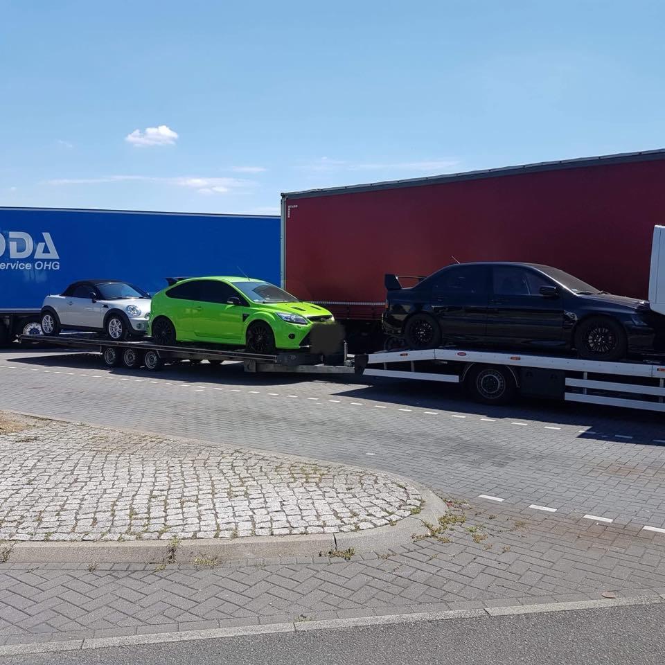 Laweta przewozi trzy sportowe samochody: srebrny kabriolet Mini Cooper, jaskrawo zielony Ford Focus RS oraz czarny Mitsubishi Lancer Evolution z dużym spojlerem, na tle błękitnego i czerwonego tira.
