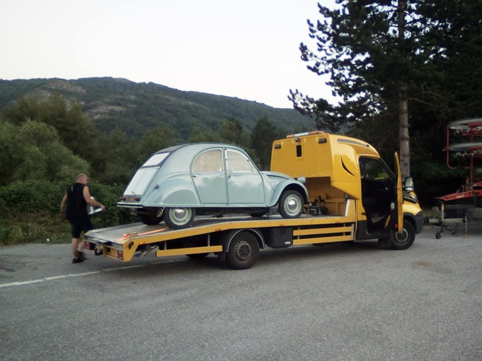 Jasnoniebieski Citroen 2CV przewożony na żółtej lawecie w scenerii górskiej, mężczyzna idzie wzdłuż lawety.