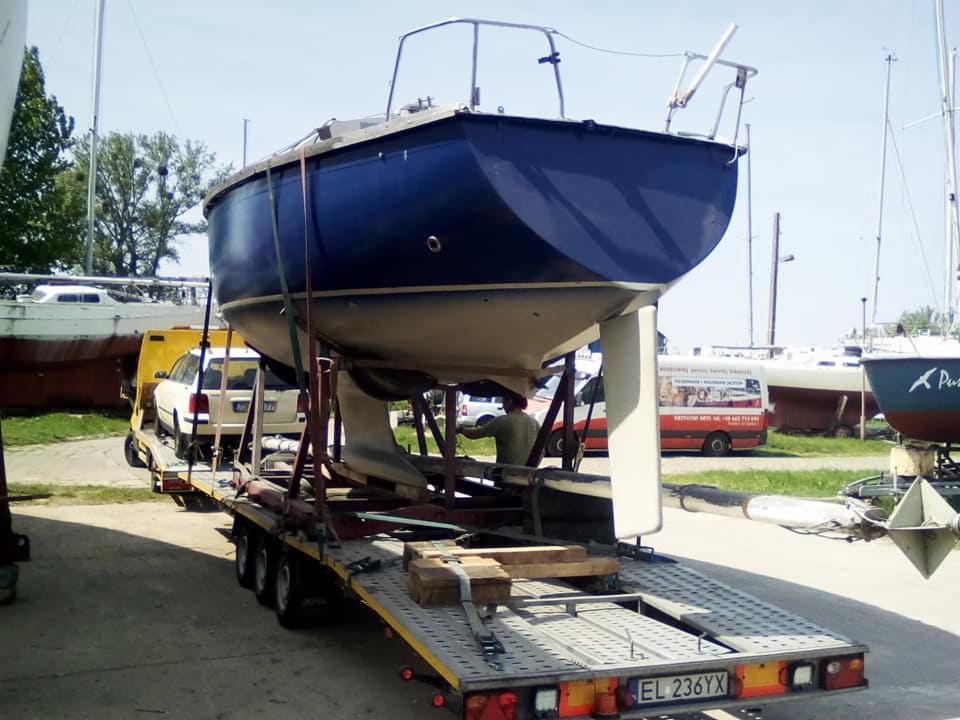 Niebieska łódź żaglowa zabezpieczona pasami na niskopodwoziowej przyczepie z żółtą lawetą w tle, widoczny numer rejestracyjny przyczepy EL 236YX.