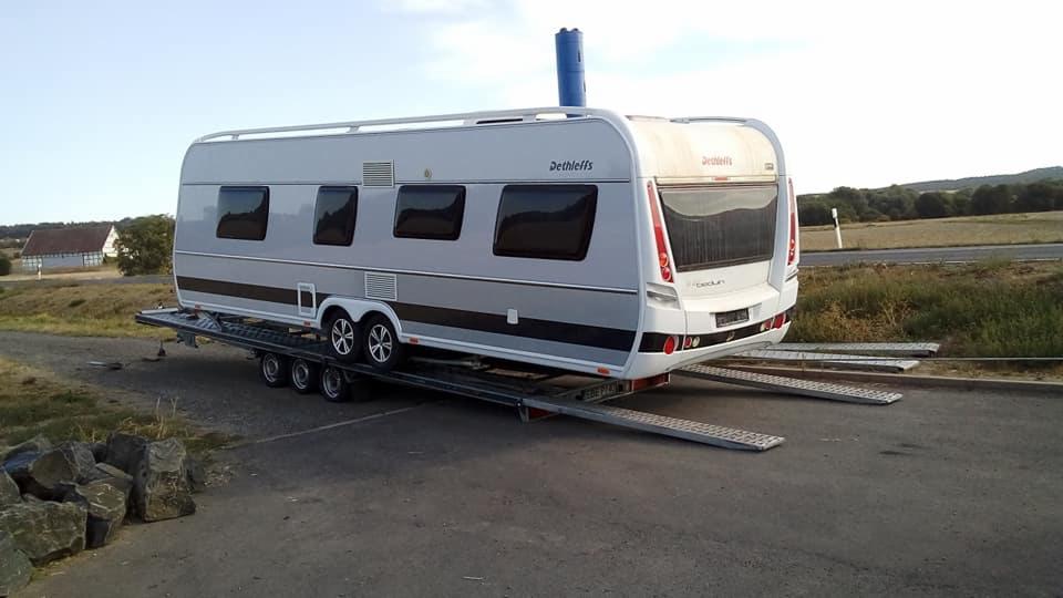 Przyczepa kempingowa marki Dethleffs, koloru szarego i białego, zabezpieczona na niskopodwoziowej platformie lawety, widok z tyłu, w tle droga i zielone pola.