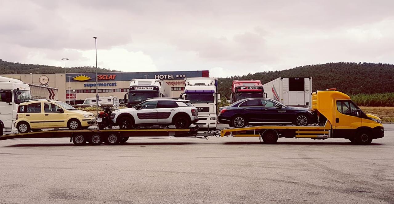 Żółta laweta z trzema samochodami osobowymi różnych marek i kolorów na platformie, zaparkowana na parkingu przed budynkiem sklepu i hotelu, w tle zielone wzgórza i pochmurne niebo.