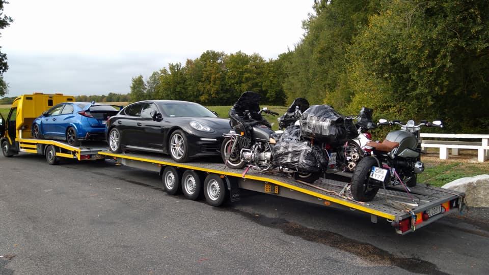 Żółta laweta z niebieskim autem osobowym, czarnym Porsche i trzema motocyklami, dwa z nich owinięte folią, na tle drzew.