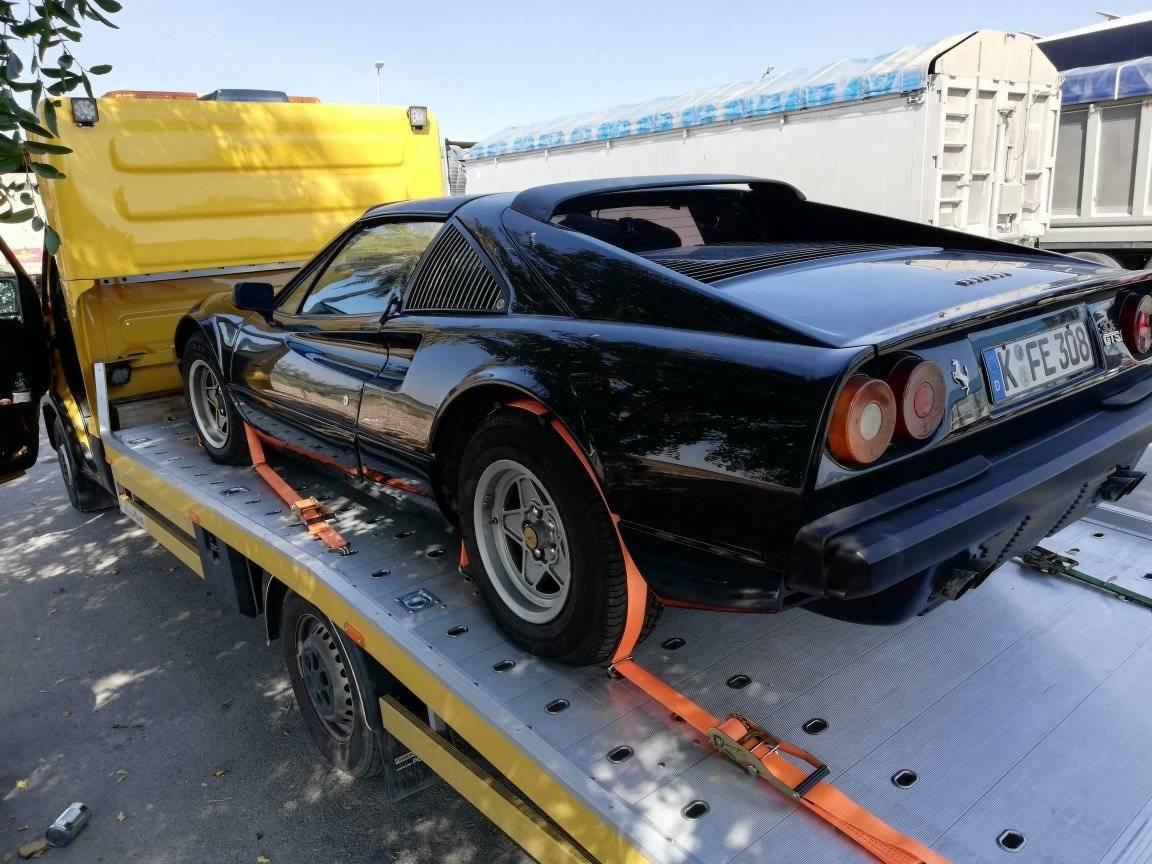 Czarny samochód sportowy Ferrari zabezpieczony pomarańczowymi pasami na żółtej lawecie, widoczny fragment niemieckiej tablicy rejestracyjnej.