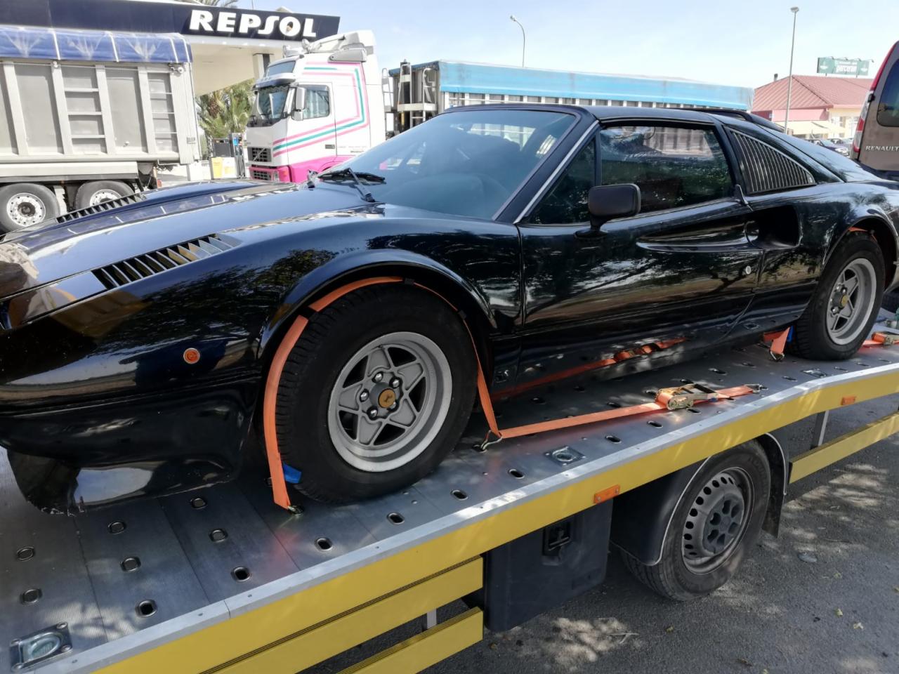 Czarny, klasyczny samochód sportowy Ferrari 308 GTS, zabezpieczony pomarańczowymi pasami, transportowany na żółtej lawecie; w tle stacja benzynowa Repsol i inne pojazdy.