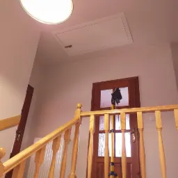 Klatka schodowa z drewnianą balustradą, widoczna klapa strychowa w suficie, oświetlenie sufitowe i lampa warsztatowa na statywie.