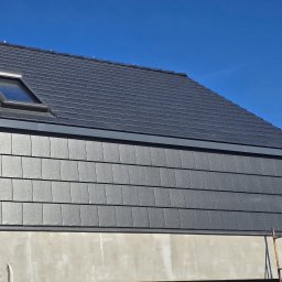 FULL BUD SPÓŁKA Z OGRANICZONĄ ODPOWIEDZIALNOŚCIĄ - Fragment dachu z czarnej dachówki bitumicznej w kształcie prostokątnych płytek, z oknem dachowym, na tle błękitnego nieba, widoczne rusztowanie.