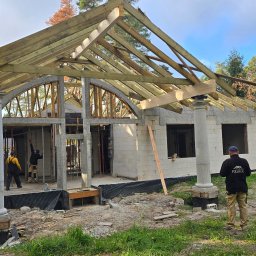 FULL BUD SPÓŁKA Z OGRANICZONĄ ODPOWIEDZIALNOŚCIĄ - Budowa domu z białych bloczków, widoczne drewniane elementy konstrukcji dachu, dwie kolumny z dekoracyjnymi głowicami, dwóch pracowników w środku budynku, trzeci obserwuje z zewnątrz, na ubraniu...