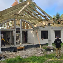 Budowa domu z białych bloczków, widoczne drewniane elementy konstrukcji dachu, dwie kolumny z dekoracyjnymi głowicami, dwóch pracowników w środku budynku, trzeci obserwuje z zewnątrz, na ubraniu...