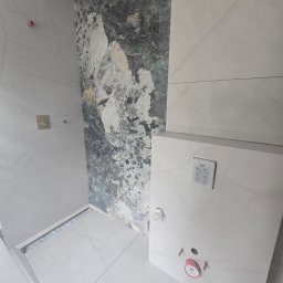 FULL BUD SPÓŁKA Z OGRANICZONĄ ODPOWIEDZIALNOŚCIĄ - Nowoczesna łazienka z jasnymi płytkami imitującymi marmur i akcentową ścianą z ciemniejszego kamienia. Widoczny liniowy odpływ prysznicowy i panel spłukujący Grohe.